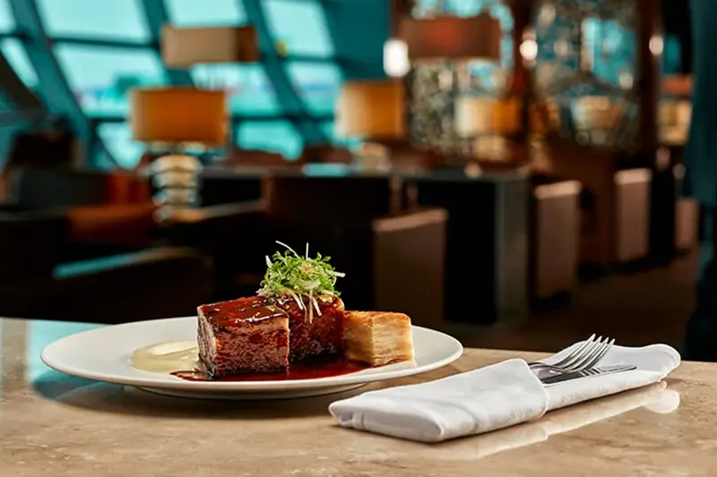 Fine dining presentation featuring rich, mahogany-colored meat resting in jus, set against a softly lit, modern lounge backdrop.