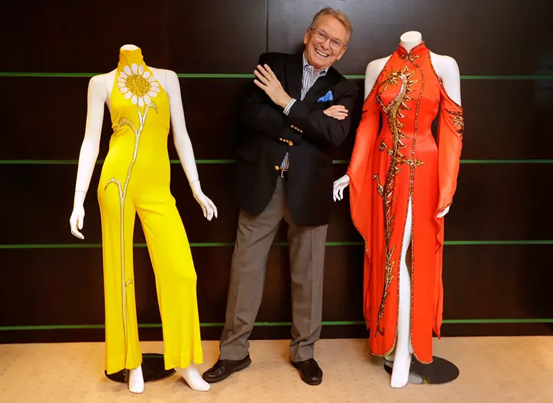 Fashion and costume designer Bob Mackie poses with two of his iconic designs, a scarlet red satin gown worn by Cher in 1975, right, and a marigold jersey jumpsuit worn by Cher between 1971-1976, left, in London on Aug. 16, 2018. (AP Photo/Kirsty Wigglesworth, File)
